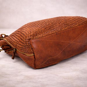 May include: A brown leather handbag with a woven top and a solid bottom. The bag has a zipper closure and a shoulder strap with a gold-colored clasp. The bag is lying on its side.