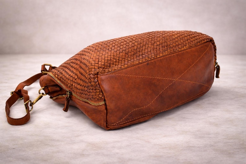 May include: A brown leather handbag with a woven top and a solid bottom. The bag has a zipper closure and a shoulder strap with a gold-colored clasp. The bag is lying on its side.