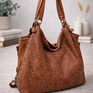 May include: A brown leather handbag with a woven design. The bag has two handles and a shoulder strap. The bag is made of brown leather and has a zipper closure. The bag is sitting on a light gray surface.