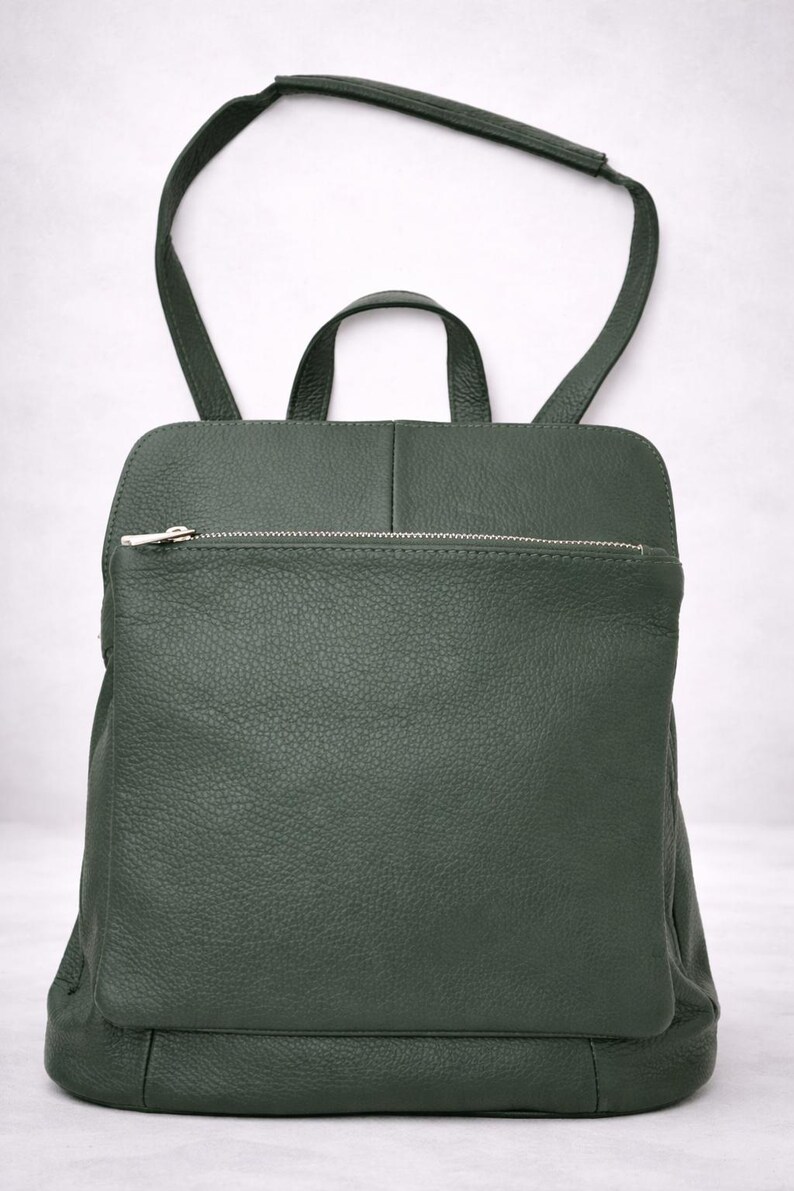 K&ouml;nnte beinhalten: Ein dunkelgr&uuml;ner Lederrucksack mit einer Rei&szlig;verschlusstasche vorne und einem oberen Griff. Der Rucksack hat verstellbare Schultergurte und einen silbernen Rei&szlig;verschluss. Die Tasche ist aus strukturiertem Leder.