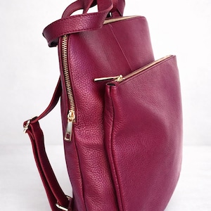 K&ouml;nnte beinhalten: Ein burgunderfarbener Lederrucksack mit goldfarbenen Rei&szlig;verschl&uuml;ssen und Beschl&auml;gen. Die Tasche hat einen oberen Griff, verstellbare Schultergurte und eine Rei&szlig;verschlusstasche vorne. Das Leder hat eine strukturierte Oberfl&auml;che.