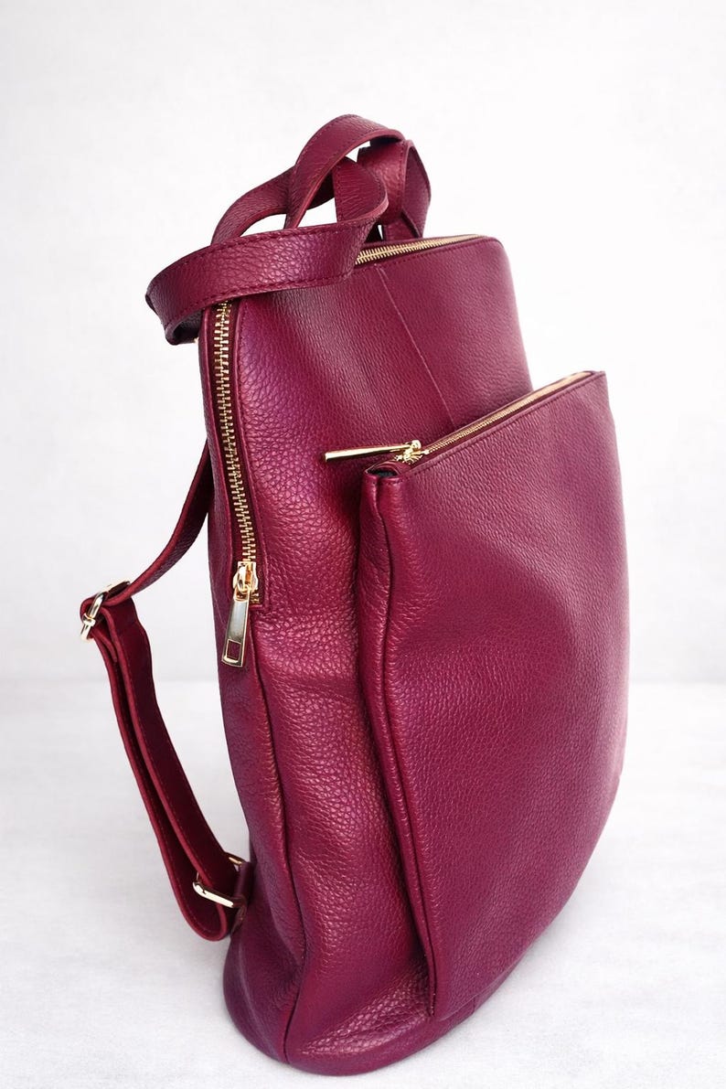 K&ouml;nnte beinhalten: Ein burgunderfarbener Lederrucksack mit goldfarbenen Rei&szlig;verschl&uuml;ssen und Beschl&auml;gen. Die Tasche hat einen oberen Griff, verstellbare Schultergurte und eine Rei&szlig;verschlusstasche vorne. Das Leder hat eine strukturierte Oberfl&auml;che.