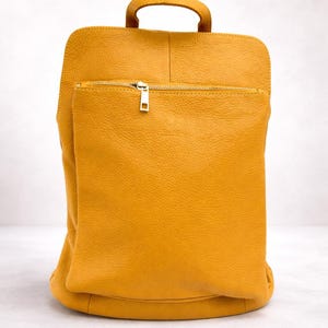 K&ouml;nnte beinhalten: Senfgelber Lederrucksack mit oberem Griff und Fronttasche mit Rei&szlig;verschluss. Die Tasche hat eine rechteckige Form mit abgerundeten Ecken und einen goldfarbenen Rei&szlig;verschluss. Das Leder hat ein strukturiertes Aussehen.