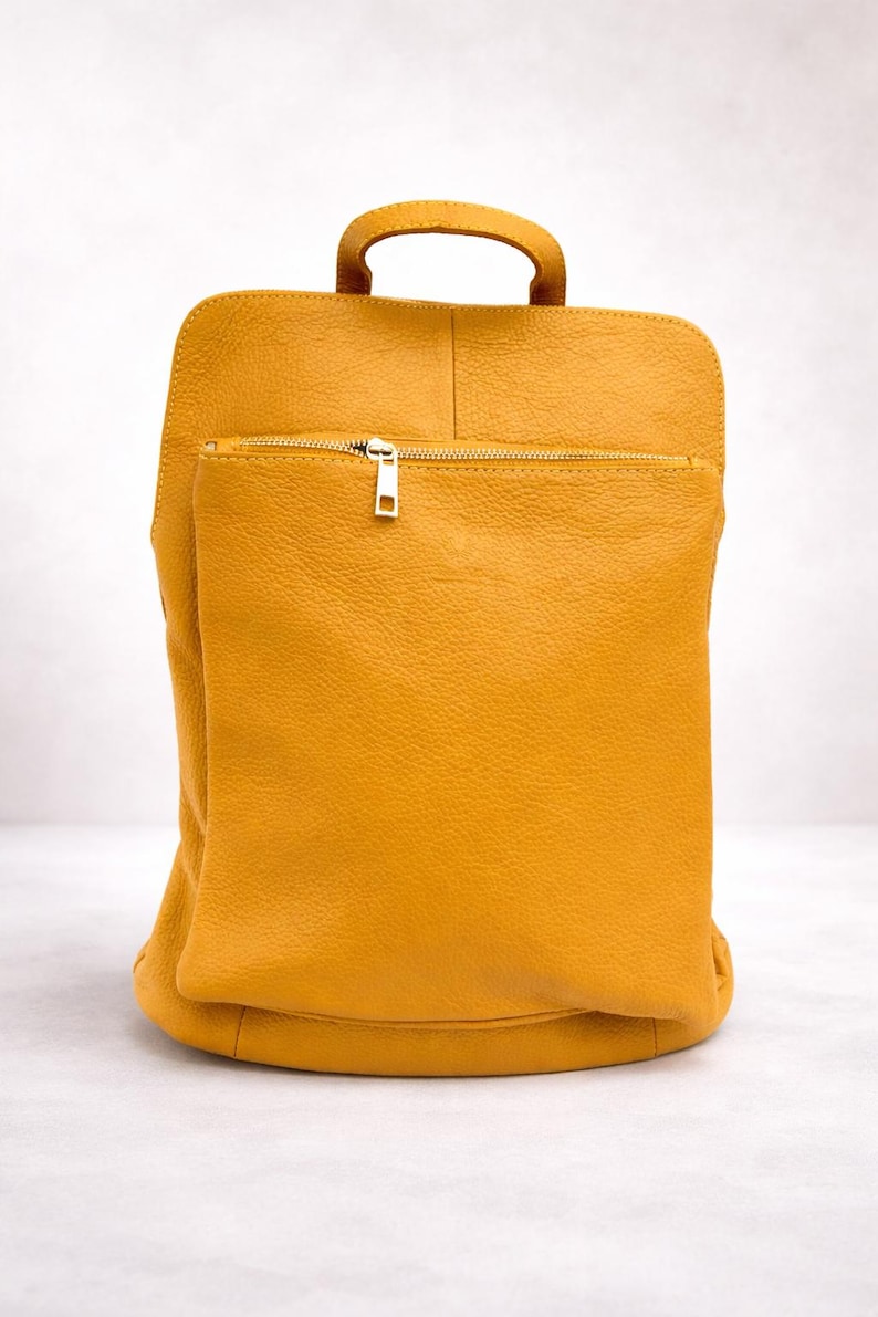 K&ouml;nnte beinhalten: Senfgelber Lederrucksack mit oberem Griff und Fronttasche mit Rei&szlig;verschluss. Die Tasche hat eine rechteckige Form mit abgerundeten Ecken und einen goldfarbenen Rei&szlig;verschluss. Das Leder hat ein strukturiertes Aussehen.