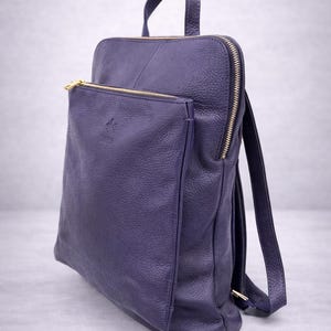 K&ouml;nnte beinhalten: Ein tiefvioletter Lederrucksack mit einer Rei&szlig;verschlusstasche vorne und goldfarbener Hardware. Die Tasche hat einen oberen Griff und verstellbare Schultergurte. Das Leder hat eine strukturierte Optik.