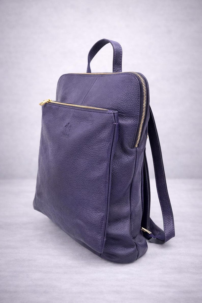 K&ouml;nnte beinhalten: Ein tiefvioletter Lederrucksack mit einer Rei&szlig;verschlusstasche vorne und goldfarbener Hardware. Die Tasche hat einen oberen Griff und verstellbare Schultergurte. Das Leder hat eine strukturierte Optik.