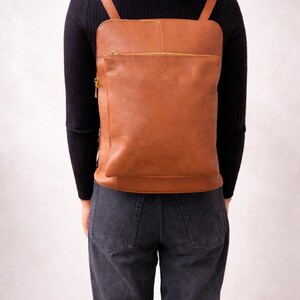 K&ouml;nnte beinhalten: Ein brauner Lederrucksack mit rechteckiger Form und einer Rei&szlig;verschlusstasche vorne. Der Rucksack hat verstellbare Schultergurte und einen goldfarbenen Rei&szlig;verschluss. Die Tasche wird auf dem R&uuml;cken getragen.