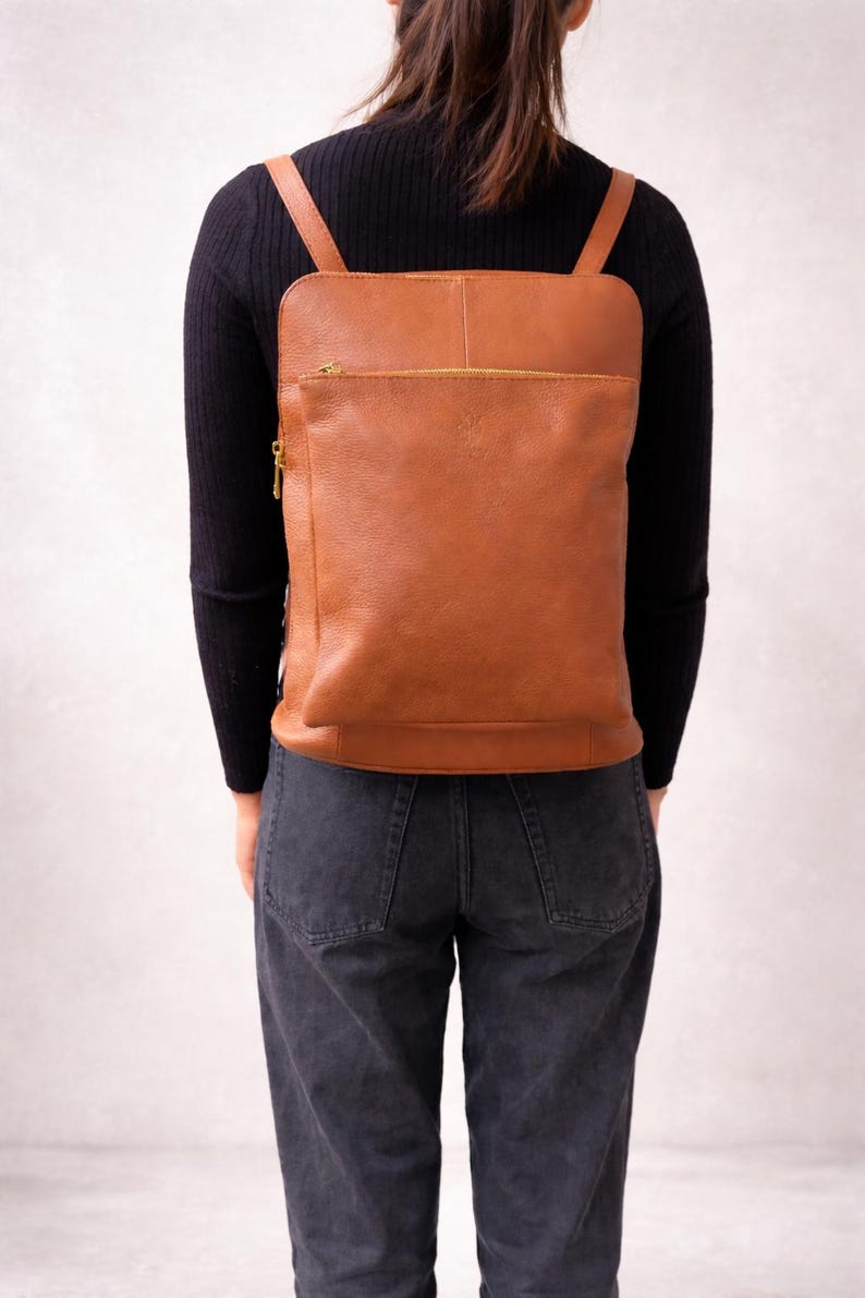 K&ouml;nnte beinhalten: Ein brauner Lederrucksack mit rechteckiger Form und einer Rei&szlig;verschlusstasche vorne. Der Rucksack hat verstellbare Schultergurte und einen goldfarbenen Rei&szlig;verschluss. Die Tasche wird auf dem R&uuml;cken getragen.