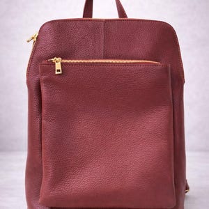 K&ouml;nnte beinhalten: Burgunderfarbener Rucksack mit einer Vordertasche und einem oberen Griff. Die Tasche hat goldfarbene Rei&szlig;verschl&uuml;sse und ein strukturiertes, lederartiges Aussehen. Das Design ist schlicht und funktional, geeignet f&uuml;r den t&auml;glichen Gebrauch.