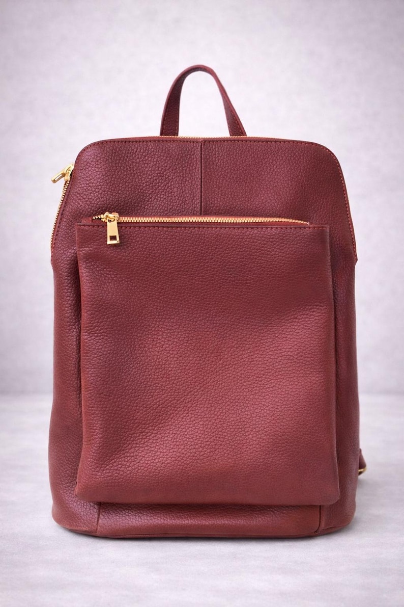 K&ouml;nnte beinhalten: Burgunderfarbener Rucksack mit einer Vordertasche und einem oberen Griff. Die Tasche hat goldfarbene Rei&szlig;verschl&uuml;sse und ein strukturiertes, lederartiges Aussehen. Das Design ist schlicht und funktional, geeignet f&uuml;r den t&auml;glichen Gebrauch.