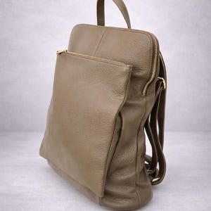 K&ouml;nnte beinhalten: Ein beigefarbener Lederrucksack mit einer Rei&szlig;verschlusstasche vorne und einem oberen Griff. Der Rucksack hat verstellbare Schultergurte und einen goldfarbenen Rei&szlig;verschluss. Das Leder hat ein strukturiertes Aussehen.