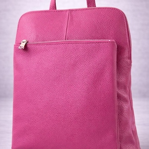 K&ouml;nnte beinhalten: Ein fuchsiafarbener Lederrucksack mit einer Rei&szlig;verschlusstasche vorne und einem oberen Griff. Der Rucksack hat eine rechteckige Form mit abgerundeten Ecken und einem silbernen Rei&szlig;verschluss. Das Leder hat eine strukturierte Oberfl&auml;che.