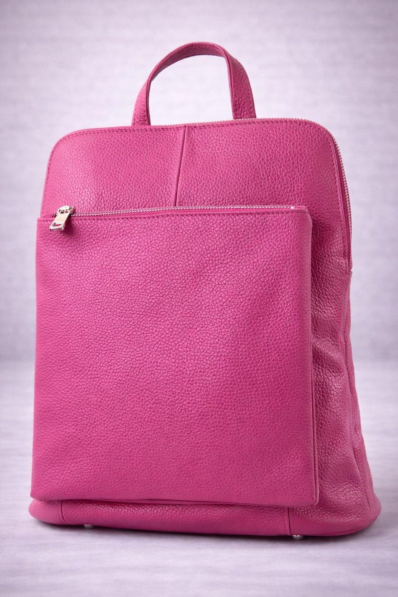 K&ouml;nnte beinhalten: Ein fuchsiafarbener Lederrucksack mit einer Rei&szlig;verschlusstasche vorne und einem oberen Griff. Der Rucksack hat eine rechteckige Form mit abgerundeten Ecken und einem silbernen Rei&szlig;verschluss. Das Leder hat eine strukturierte Oberfl&auml;che.