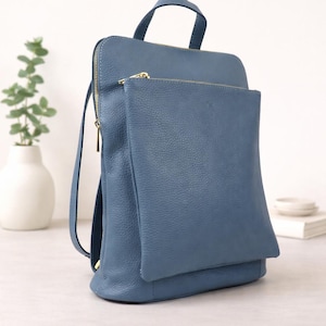 K&ouml;nnte beinhalten: Ein blauer Lederrucksack mit passender Vordertasche. Der Rucksack hat einen oberen Griff, verstellbare Tr&auml;ger und einen goldenen Rei&szlig;verschluss. Eine wei&szlig;e Vase mit Gr&uuml;nzeug befindet sich im Hintergrund.
