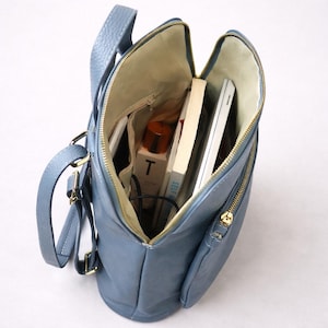 K&ouml;nnte beinhalten: Offene Ansicht eines blauen Lederrucksacks mit goldenen Rei&szlig;verschl&uuml;ssen, der seinen Inhalt zeigt. Im Inneren befinden sich B&uuml;cher, ein wei&szlig;er Laptop, eine Flasche mit dem Buchstaben "T" und andere Gegenst&auml;nde. Die Tasche hat einen oberen Griff und verstellbare Tr&auml;ger.