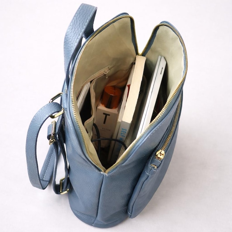 K&ouml;nnte beinhalten: Offene Ansicht eines blauen Lederrucksacks mit goldenen Rei&szlig;verschl&uuml;ssen, der seinen Inhalt zeigt. Im Inneren befinden sich B&uuml;cher, ein wei&szlig;er Laptop, eine Flasche mit dem Buchstaben "T" und andere Gegenst&auml;nde. Die Tasche hat einen oberen Griff und verstellbare Tr&auml;ger.