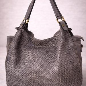 May include: A gray leather handbag with a woven design. The bag has two handles and a zipper closure. The hardware is gold-colored. The bag is a neutral color and appears to be made of high-quality leather.