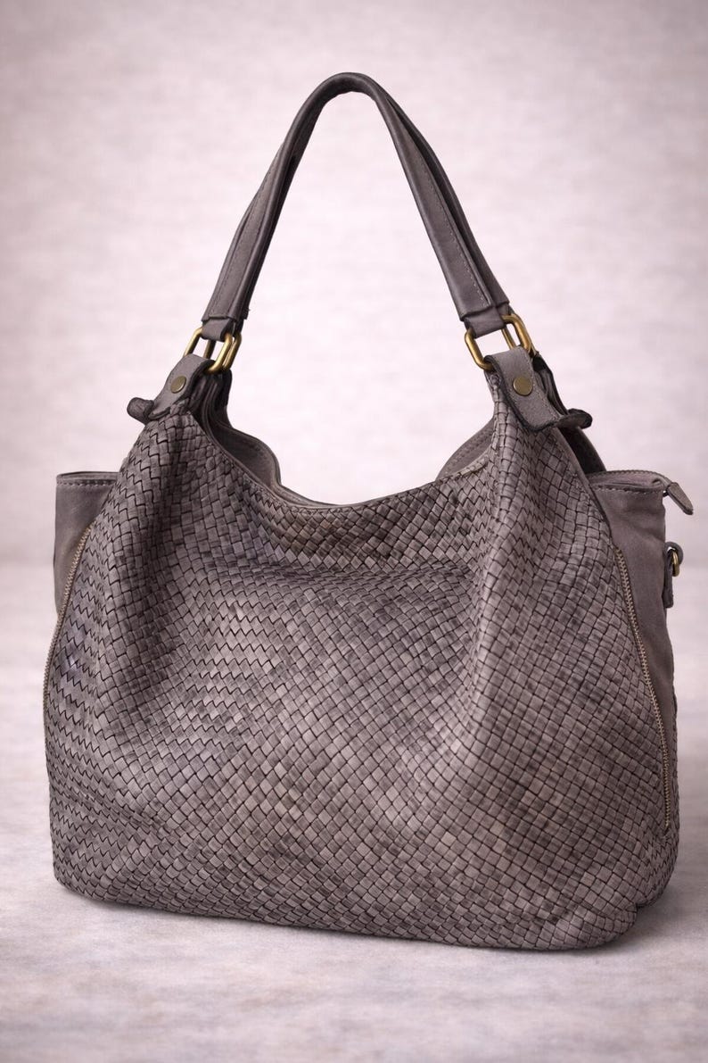 May include: A gray leather handbag with a woven design. The bag has two handles and a zipper closure. The hardware is gold-colored. The bag is a neutral color and appears to be made of high-quality leather.
