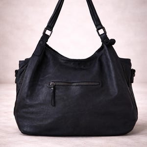 May include: Black leather shoulder bag with a zippered front pocket and two top handles. The bag has a slouchy design and a zipper closure. The bag is made of a soft, textured leather.