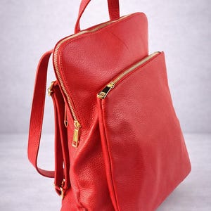 K&ouml;nnte beinhalten: Ein leuchtend roter Rucksack aus strukturiertem Leder. Er hat einen oberen Griff, verstellbare Tr&auml;ger und eine Vordertasche. Goldfarbene Rei&szlig;verschl&uuml;sse und Beschl&auml;ge bilden einen Kontrast. Der Rucksack ist f&uuml;r den t&auml;glichen Gebrauch konzipiert.