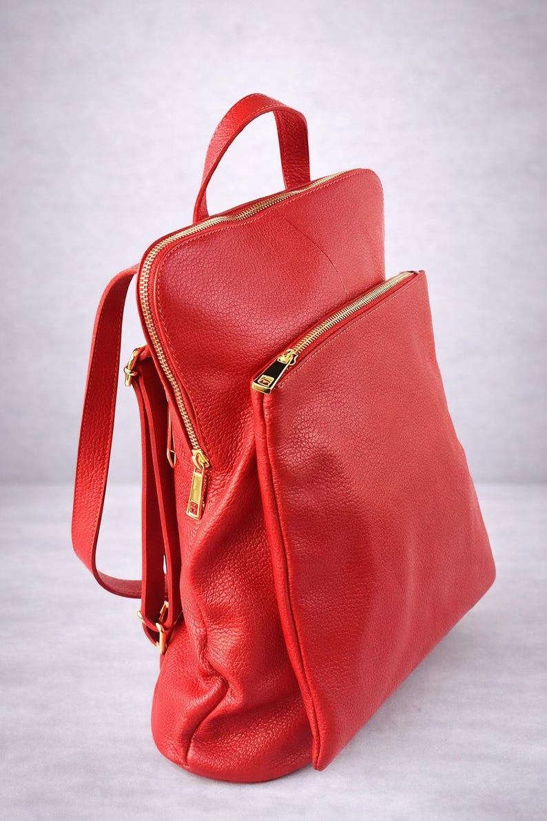 K&ouml;nnte beinhalten: Ein leuchtend roter Rucksack aus strukturiertem Leder. Er hat einen oberen Griff, verstellbare Tr&auml;ger und eine Vordertasche. Goldfarbene Rei&szlig;verschl&uuml;sse und Beschl&auml;ge bilden einen Kontrast. Der Rucksack ist f&uuml;r den t&auml;glichen Gebrauch konzipiert.