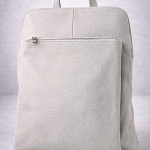 K&ouml;nnte beinhalten: Ein hellgrauer Lederrucksack mit einem oberen Griff und einer Rei&szlig;verschlusstasche vorne. Der Rucksack hat eine rechteckige Form mit abgerundeten Ecken und einer strukturierten Oberfl&auml;che. Die Rei&szlig;verschlussschieber sind silberfarben.