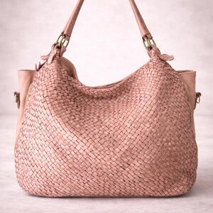 May include: A blush-colored woven leather handbag with a curved top and two handles. The bag features a large, open main compartment and a zippered pocket on the side. The handles are attached with gold-colored metal hardware.
