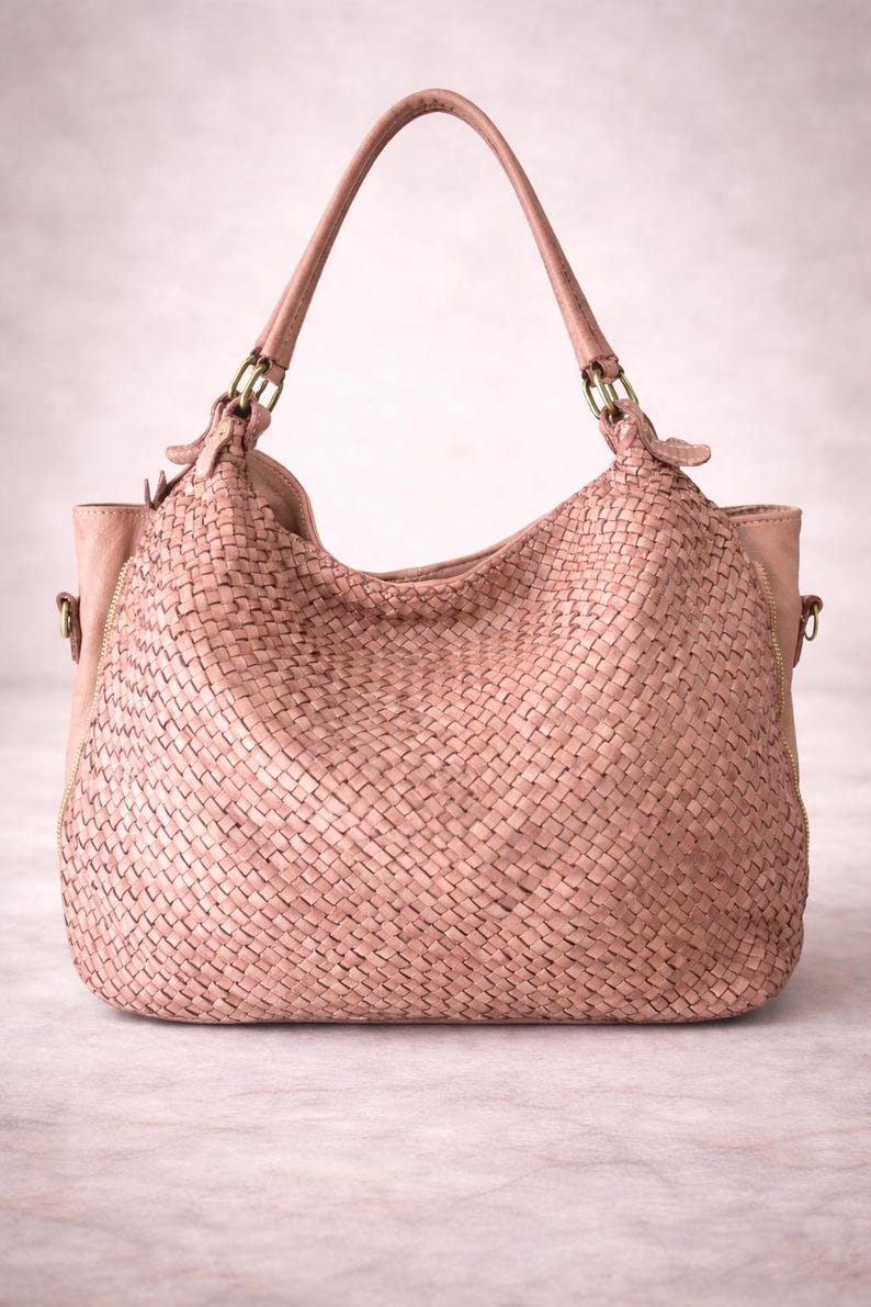 May include: A blush-colored woven leather handbag with a curved top and two handles. The bag features a large, open main compartment and a zippered pocket on the side. The handles are attached with gold-colored metal hardware.