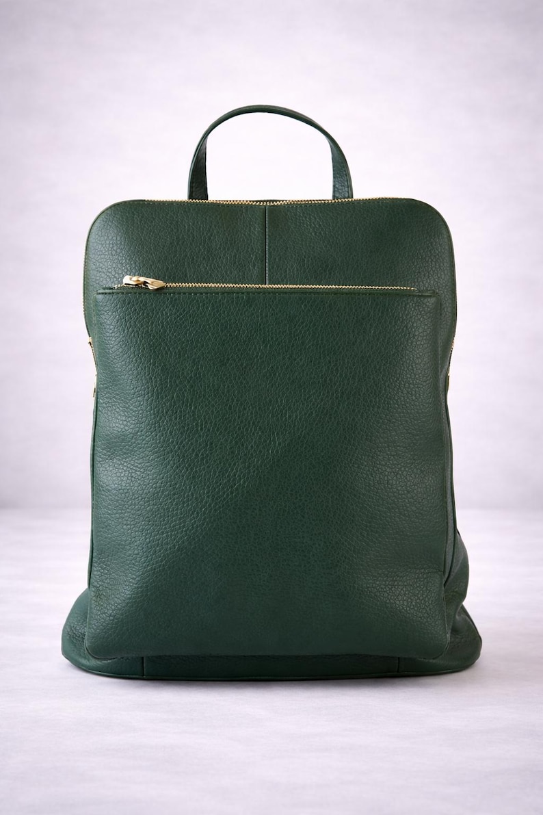 K&ouml;nnte beinhalten: Ein dunkelgr&uuml;ner Lederrucksack mit einem oberen Griff und einer Rei&szlig;verschlusstasche vorne. Der Rucksack hat einen goldfarbenen Rei&szlig;verschluss und eine strukturierte Oberfl&auml;che. Die Tasche hat eine rechteckige Form.