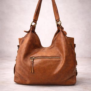 May include: A brown leather handbag with two handles and a zippered front pocket. The bag has a slouchy design and brass-colored hardware. The leather has a distressed look, giving it a vintage feel. The bag is suitable for everyday use.