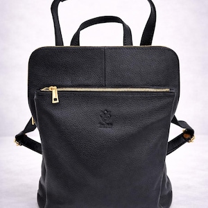 K&ouml;nnte beinhalten: Schwarzer Lederrucksack mit Tragegriff und verstellbaren Schultergurten. Der Rucksack hat eine Rei&szlig;verschlusstasche vorne und goldfarbene Hardware. Die Tasche hat eine strukturierte Oberfl&auml;che und ein kleines Logo auf der Vorderseite.
