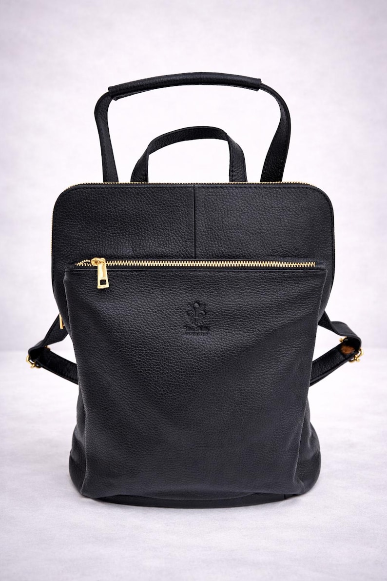 K&ouml;nnte beinhalten: Schwarzer Lederrucksack mit Tragegriff und verstellbaren Schultergurten. Der Rucksack hat eine Rei&szlig;verschlusstasche vorne und goldfarbene Hardware. Die Tasche hat eine strukturierte Oberfl&auml;che und ein kleines Logo auf der Vorderseite.