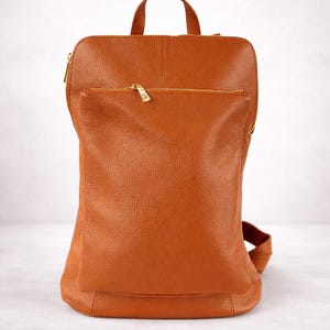 K&ouml;nnte beinhalten: Ein brauner Lederrucksack mit Tragegriff und verstellbaren Tr&auml;gern. Der Rucksack hat eine Rei&szlig;verschlusstasche vorne und einen goldfarbenen Rei&szlig;verschluss. Die Tasche ist aus strukturiertem Leder.