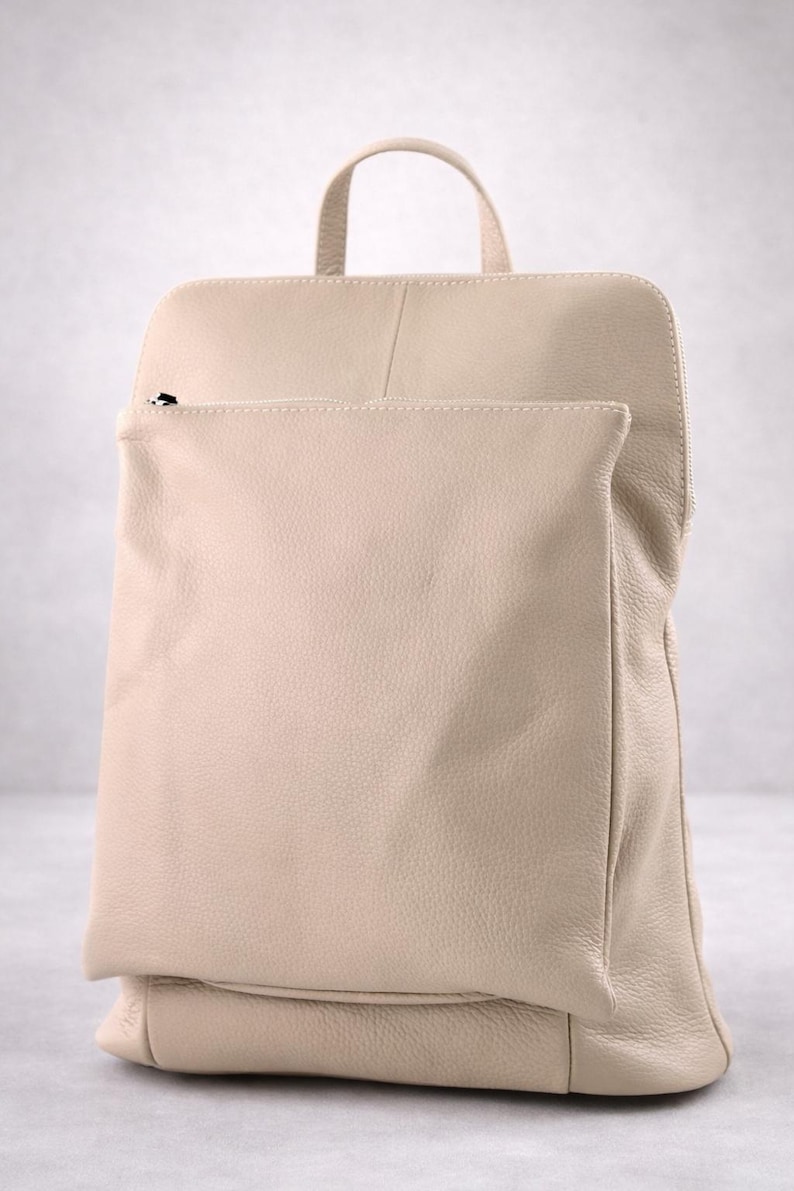 K&ouml;nnte beinhalten: Beigefarbener Lederrucksack mit oberem Griff und Vordertasche. Die Tasche hat eine rechteckige Form mit abgerundeten Ecken und einen Rei&szlig;verschluss an der Vordertasche. Das Leder hat ein strukturiertes Aussehen.