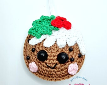 May include: A crocheted Christmas pudding ornament with a white icing top, green holly leaves, and red berries. The pudding has a smiling face with black eyes and brown buttons. The ornament is hanging from a white ribbon. The text "Sam's Yarn Barn Crochet Magic" is visible in the bottom right corner.