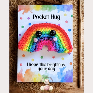 Puede incluir: Un arcoíris de ganchillo hecho a mano con ojos de botón negros y mejillas de cuentas rosas, adjunto a una tarjeta con el texto "Pocket Hug" y "I hope this brightens your day". La tarjeta tiene un fondo de acuarela.