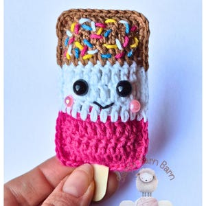 May include: A crocheted popsicle toy with a brown chocolate top, white vanilla middle, and pink strawberry bottom. The popsicle has a cute face with black eyes and pink cheeks. It is held by a hand and has a wooden stick.