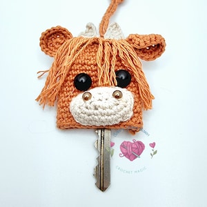 May include: A crocheted highland cow keychain with brown and white fur, black eyes, and a gold nose. The keychain is attached to a silver key.
