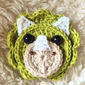May include: A handmade crochet Triceratops dinosaur head in shades of green, white, and beige. The dinosaur has large black eyes and small brown bead accents. The item is likely a decorative item or a toy.