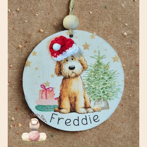 Op de afbeelding: Ronde houten kerstversiering met een lichtbruine hond met een rood-witte kerstmuts. De versiering heeft een kerstboom, cadeaus en de naam "Freddie" erop. Een houten kraal en touw zijn bevestigd om op te hangen.