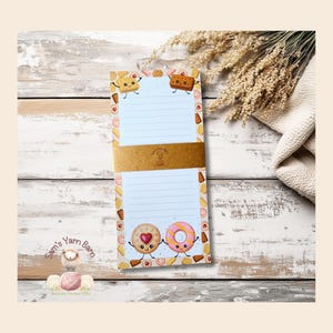 May include: A notepad with a fun design of cartoon cookies and pastries. The notepad has lined pages and a brown paper band. The design includes a smiling cookie with a heart and a donut with sprinkles. The notepad is on a white wooden surface.