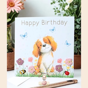 Könnte beinhalten: Geburtstagskarte mit der Aufschrift "Happy Birthday". Die Karte zeigt eine Cartoon-Illustration eines Hundes mit orange-weißem Fell, umgeben von Blumen, Schmetterlingen und Insekten.