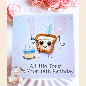 May include: A handmade birthday card featuring a crocheted toast character holding pink balloons shaped as the numbers 1 and 8. A small blue cake with candles is on the left. The text reads "A Little Toast To Your 18th Birthday."