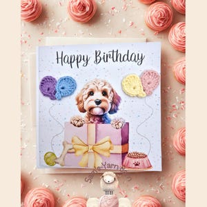 Könnte beinhalten: Eine Geburtstagskarte mit einem süßen Hund in einer Geschenkbox mit einer gelben Schleife. Die Karte hat einen weißen Hintergrund mit Konfetti und den Worten "Happy Birthday" oben. Es gibt drei gehäkelte Ballons in den Farben Rosa, Blau und Gelb.