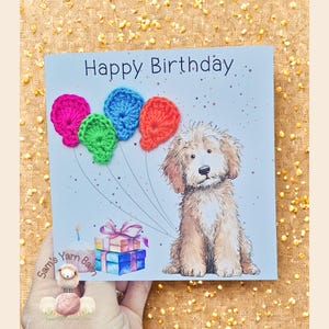 May include: A birthday card with the words "Happy Birthday" at the top. The card features a watercolor illustration of a fluffy dog and crocheted balloons in pink, blue, green, and orange. A gift and a candle are also depicted.