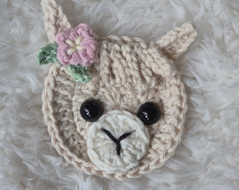 May include: A crocheted llama face with black button eyes, a white muzzle, and a pink flower with green leaves. The llama is cream-colored and has small, pointed ears. The craft item is likely a patch or applique.