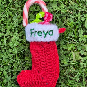 May include: A red crocheted stocking with a white cuff and the name "Freyja" embroidered on it. The stocking is filled with a candy cane and other treats.