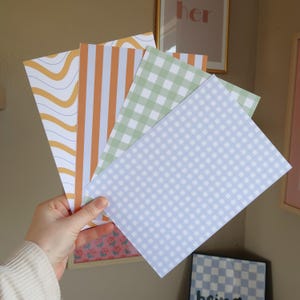 Op de afbeelding: Een hand houdt een waaier van papieren vellen met patronen vast. De ontwerpen omvatten golvende lijnen, verticale strepen, ruitjes en een dambordpatroon. De kleuren zijn oranje, wit, groen en blauw. De papieren zijn geschikt voor briefpapier of knutselprojecten.