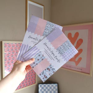 May include: Two "Romantic Paper Bundle" packs with patterned paper in pink, blue, and purple. Each pack contains eight A5 journal papers. The packs are held in front of framed artwork on a neutral wall.