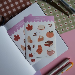 Op de afbeelding: Twee stickervellen met een gezellig herfstthema, met illustraties van bladeren, pompoenen, kaarsen en boeken. De vellen hebben een witte achtergrond met een roze rand en de tekst "COZY FALL STICKER SHEET".
